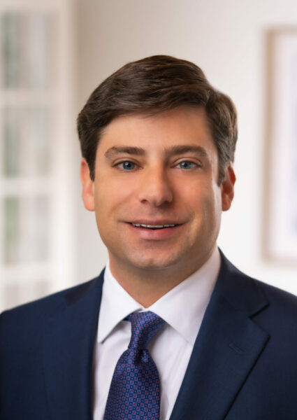 Man in a navy suit, white shirt, and patterned tie, smiling at the camera in a well-lit, indoor setting with a blurred background, embodying the professionalism of a full service law firm.