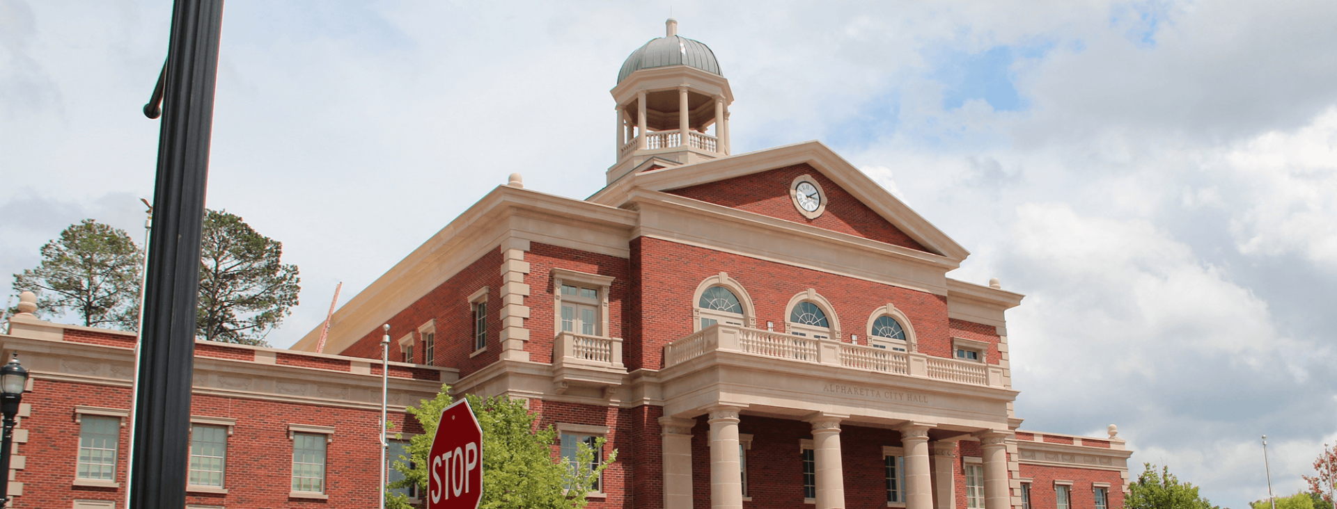 Red brick courthouse with columns, a central clock, and a domed cupola, with a stop sign in the foreground and partly cloudy skies overhead.