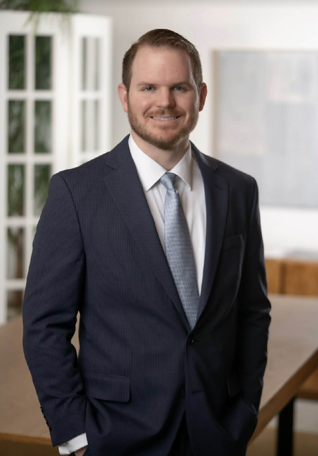 Man in a dark suit and light blue tie stands indoors, smiling, with hands in pockets. Office setting of a full service law firm with desk and glass doors in the background.