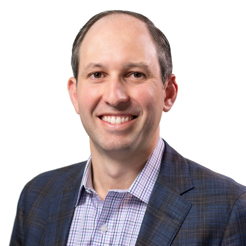 A man with short brown hair, wearing a plaid suit jacket and a checkered dress shirt, smiles at the camera against a plain white background, embodying the approachable professionalism of a full service law firm.