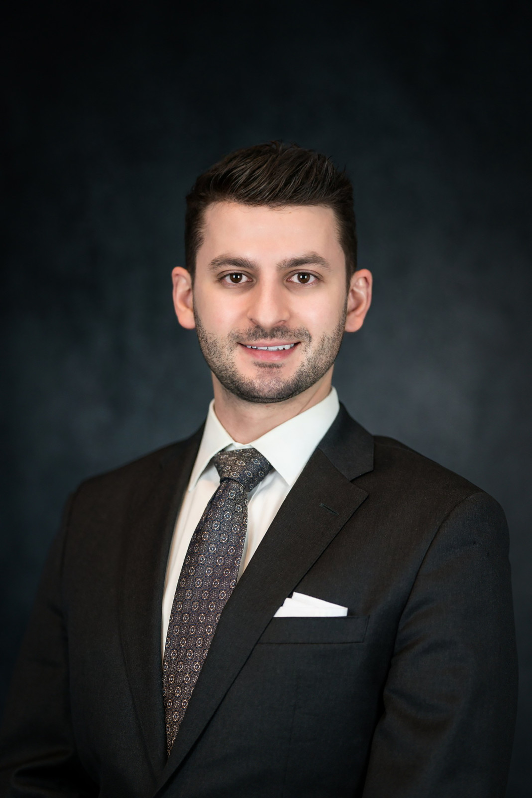 A man in a dark suit and patterned tie stands in front of a dark, textured background, looking at the camera with a slight smile—exuding the professionalism and confidence you’d expect from an attorney at a full service law firm.