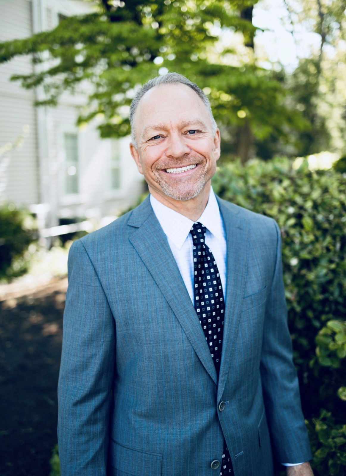 A man in a blue suit and patterned tie stands outside near greenery, smiling at the camera, embodying the professionalism often seen among corporate and business law attorneys.