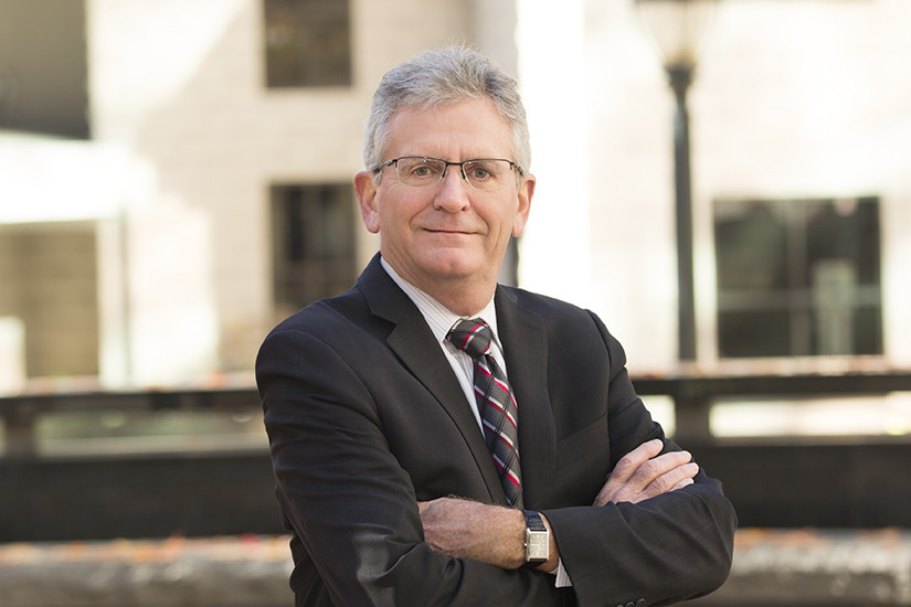 A man in a suit and tie with gray hair and glasses stands outdoors with his arms crossed, in front of a blurred building background, portraying the confidence and professionalism often associated with corporate and business law attorneys.