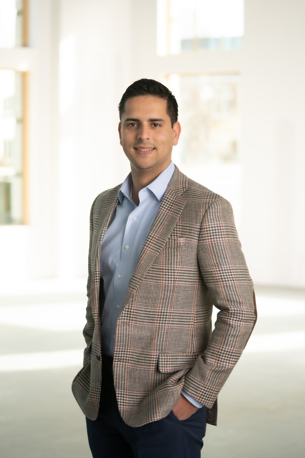 Man in a plaid blazer and blue shirt stands in a bright room with large windows, smiling with one hand in his pocket—a confident presence that reflects the professionalism of top corporate and business law attorneys.
