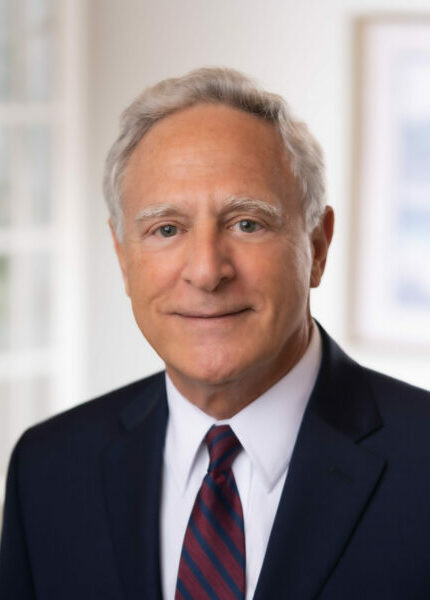 An older man with gray hair wearing a dark suit, white shirt, and striped tie is smiling slightly in a well-lit, indoor setting, embodying the professionalism associated with a full service law firm.