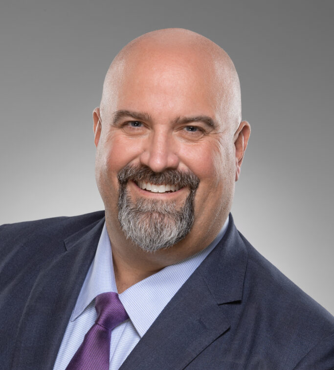 A bald man with a goatee and mustache smiles, wearing a dark suit, light blue shirt, and purple tie, in front of a plain gray background—presenting the approachable yet professional demeanor often associated with corporate and business law attorneys.