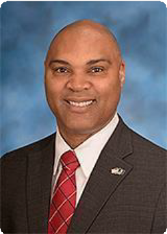 A man in a dark suit, white shirt, and red patterned tie smiles in a formal portrait against a blue background, reflecting the professional image often associated with corporate and business law attorneys.
