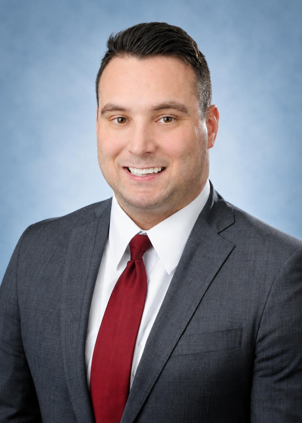 Man in a gray suit, white shirt, and red tie smiling at the camera against a light blue background.