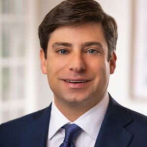 Man in a navy suit, white shirt, and patterned tie, smiling at the camera in a well-lit, indoor setting with a blurred background, embodying the professionalism of a full service law firm.