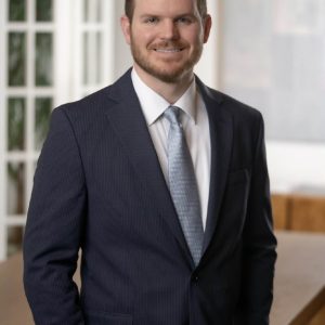 Man in a dark suit and light blue tie stands indoors, smiling, with hands in pockets. Office setting of a full service law firm with desk and glass doors in the background.
