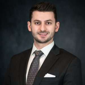 A man in a dark suit and patterned tie stands in front of a dark, textured background, looking at the camera with a slight smile—exuding the professionalism and confidence you’d expect from an attorney at a full service law firm.