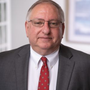 A man wearing glasses, a gray suit, white shirt, and red patterned tie stands indoors in front of a blurred background, embodying the professional image of a full service law firm.