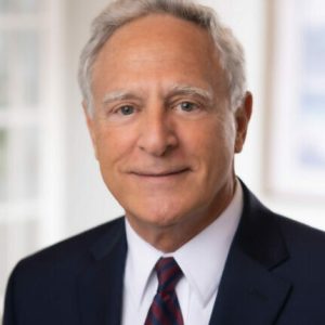 An older man with gray hair wearing a dark suit, white shirt, and striped tie is smiling slightly in a well-lit, indoor setting, embodying the professionalism associated with a full service law firm.